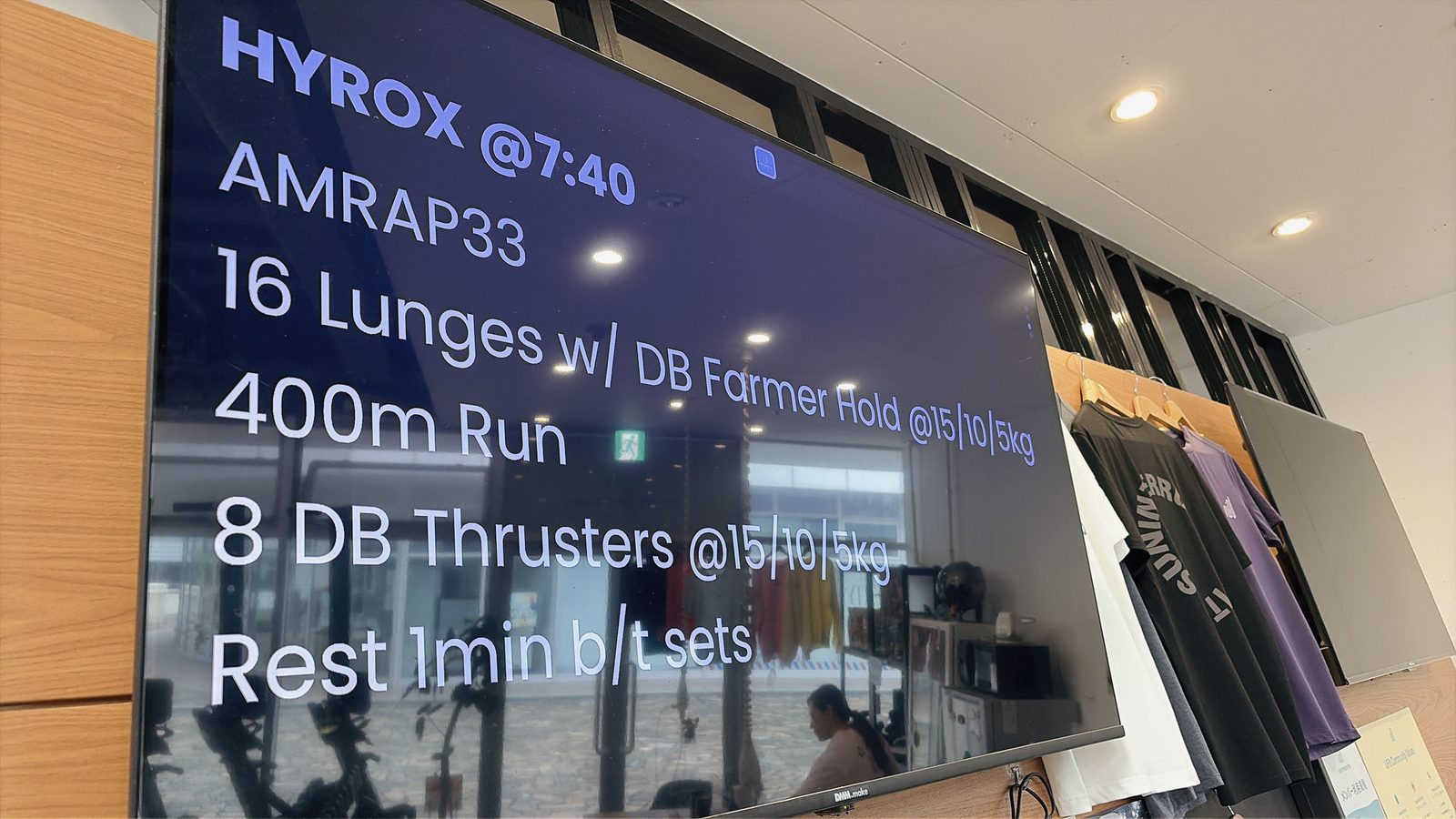 ジムのモニターに表示されたHYROXクラスのメニュー。AMRAP33、16 Lunges w/ DB Farmer Hold、400m Run、8 DB Thrusters