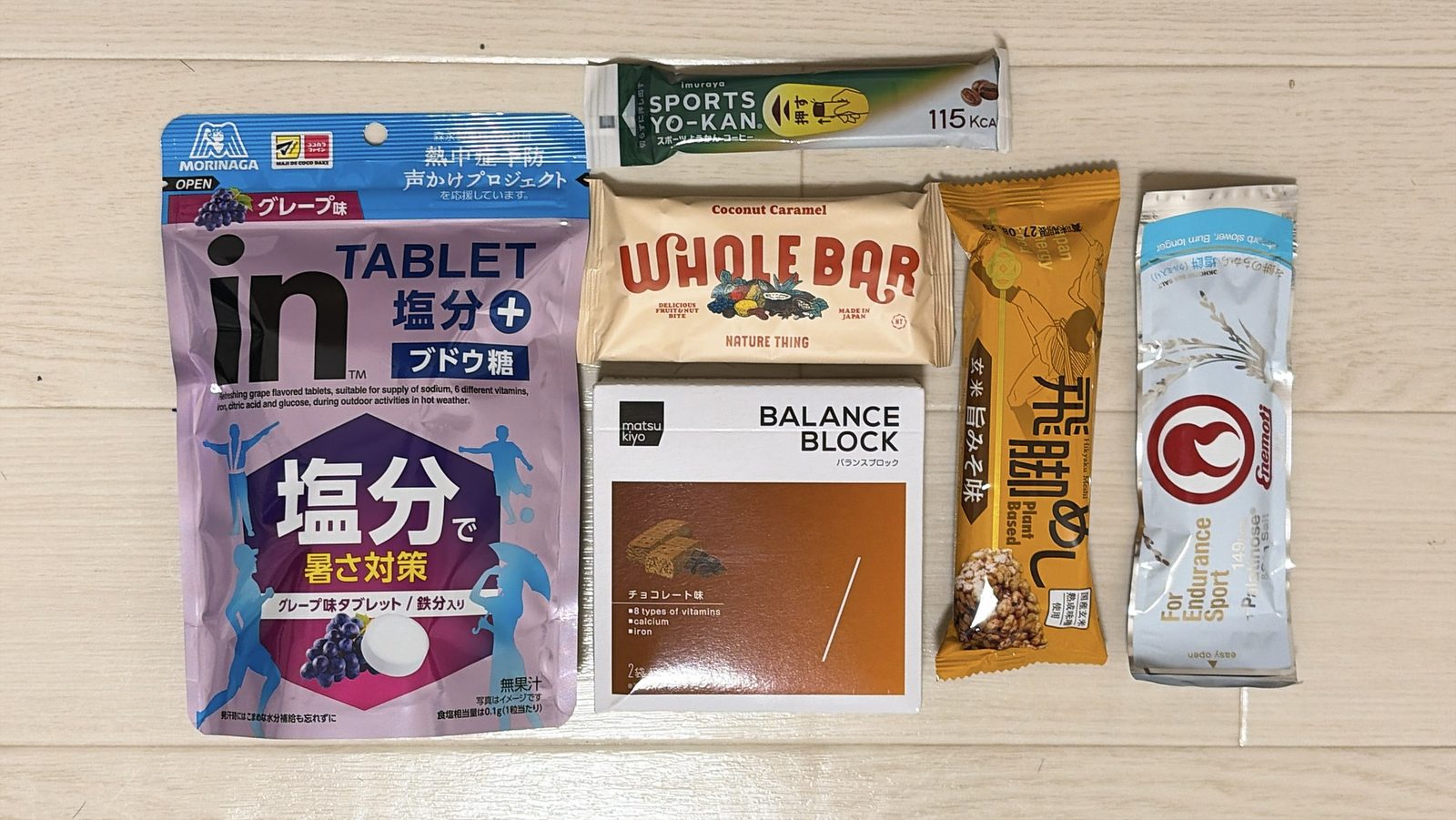 床に並べた携帯食6つ。森永製菓 inタブレット 塩分プラス グレープ味、井村屋 スポーツようかん コーヒー、NATURE THING WHOLE BAR ココナッツキャラメル、マツキヨ バランスブロック チョコレート味、飛脚めし 玄米 旨みそ味、Enemoti 塩餅