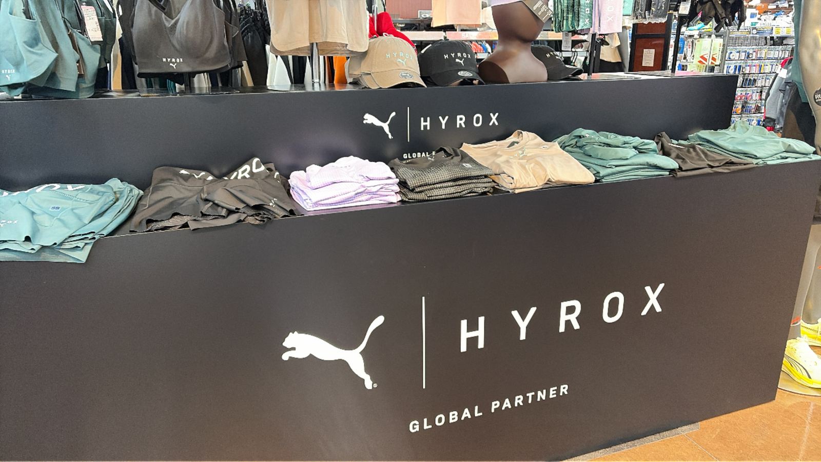 スポーツショップの売り場。黒い什器に PUMA と HYROX のロゴ、GLOBAL PARTNER の文字。上にウェアと帽子が並んでいる