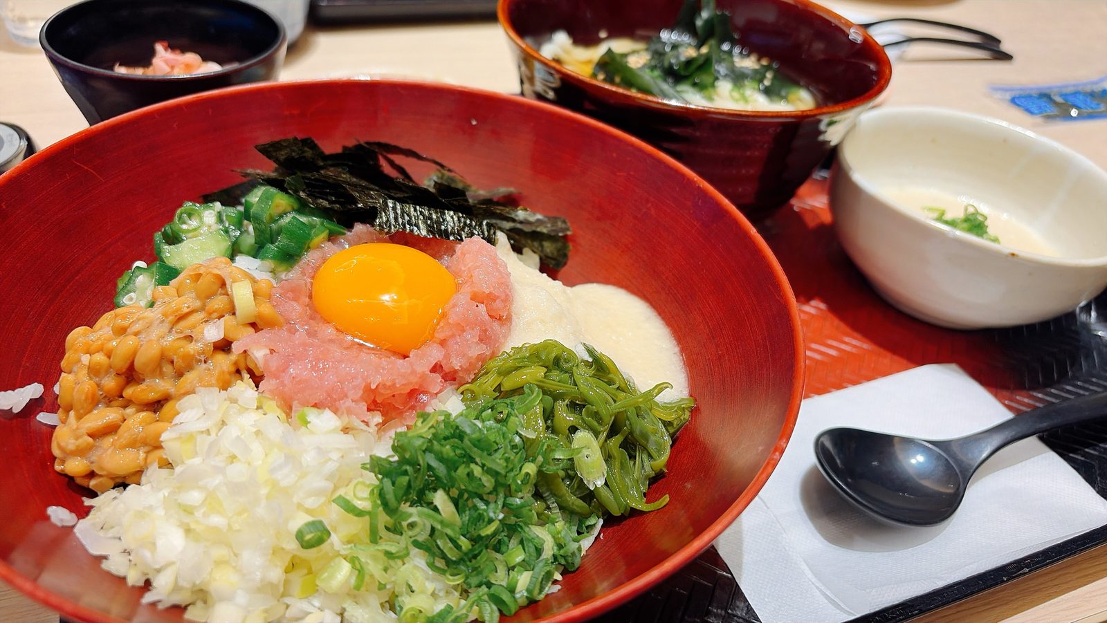 大戸屋ばくだん丼定食。納豆、まぐろ、卵黄、とろろ、オクラ、めかぶ、ねぎ。奥にわかめのうどん