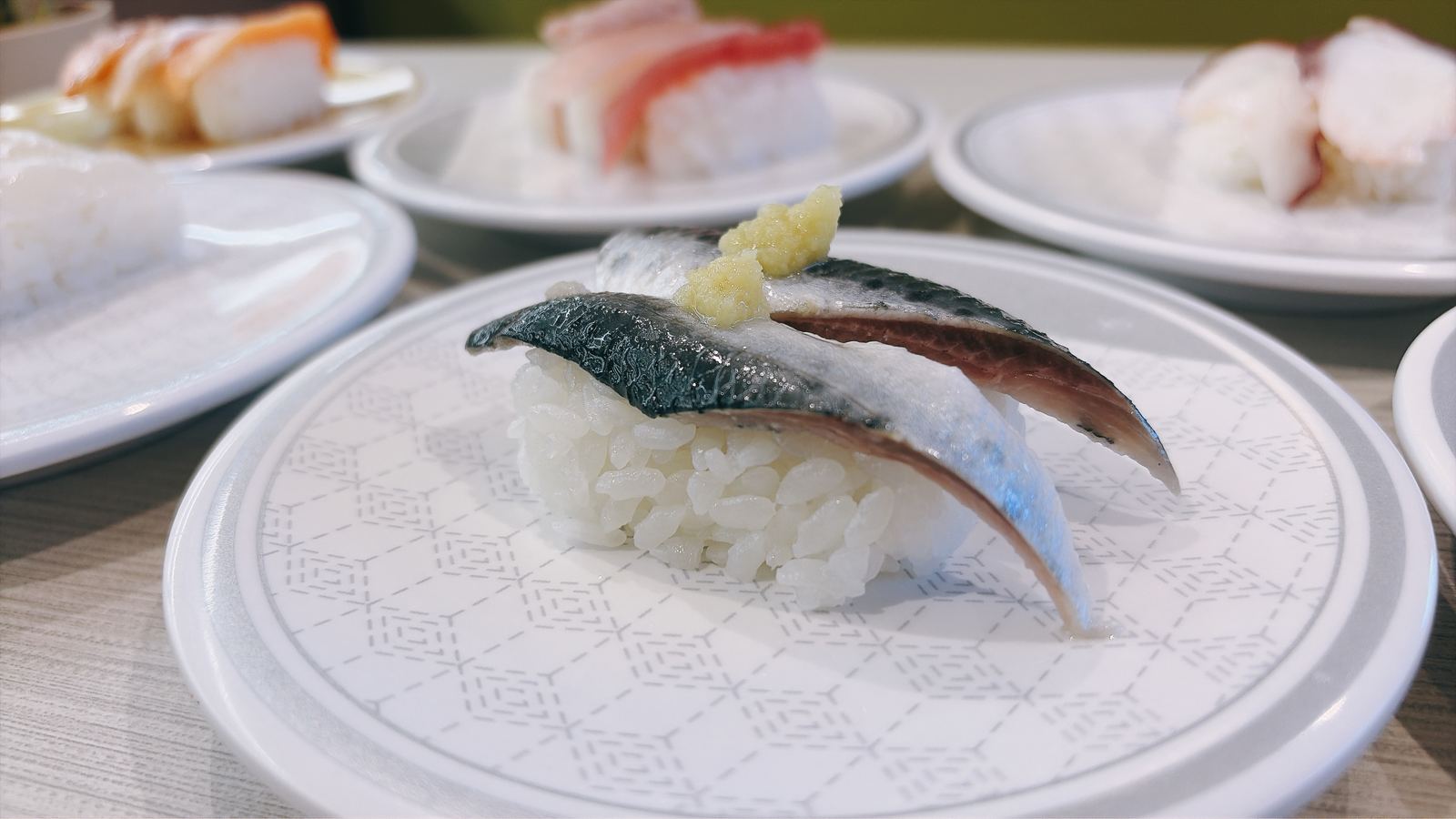 回転寿司の皿にのったサバの握り。おろし生姜がのっている。奥にも数皿並んでいる